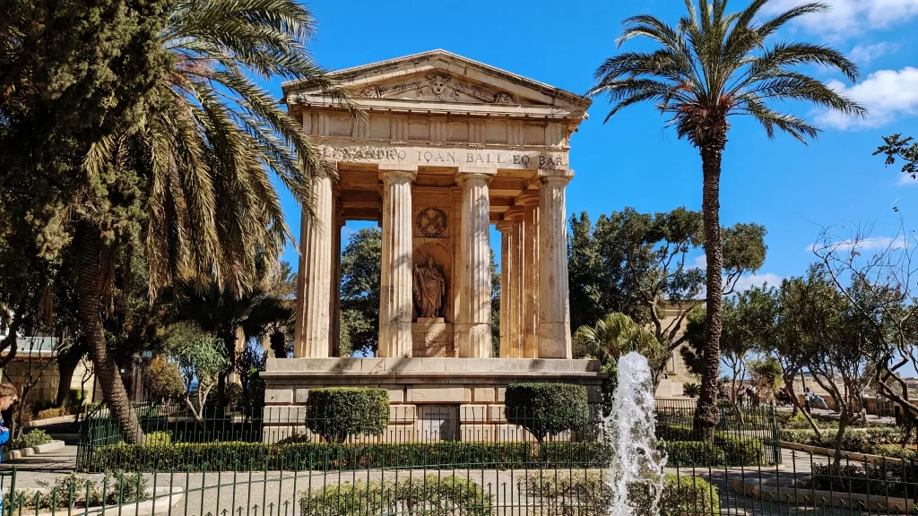A Roman style stone structure with columns and a fountain in front of it. Palm trees fame the structure. Lower Barrakka gardens is one of the best things to do in Malta.