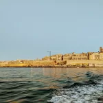 Explore the coastal cityscape of Malta, where historic stone buildings bask in the golden glow of sunset. In this serene travel guide scene, gentle waves ripple across the calm sea in the foreground, evoking a sense of wanderlust and inviting you to discover more things to do.