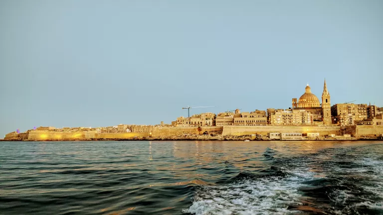 Explore the coastal cityscape of Malta, where historic stone buildings bask in the golden glow of sunset. In this serene travel guide scene, gentle waves ripple across the calm sea in the foreground, evoking a sense of wanderlust and inviting you to discover more things to do.