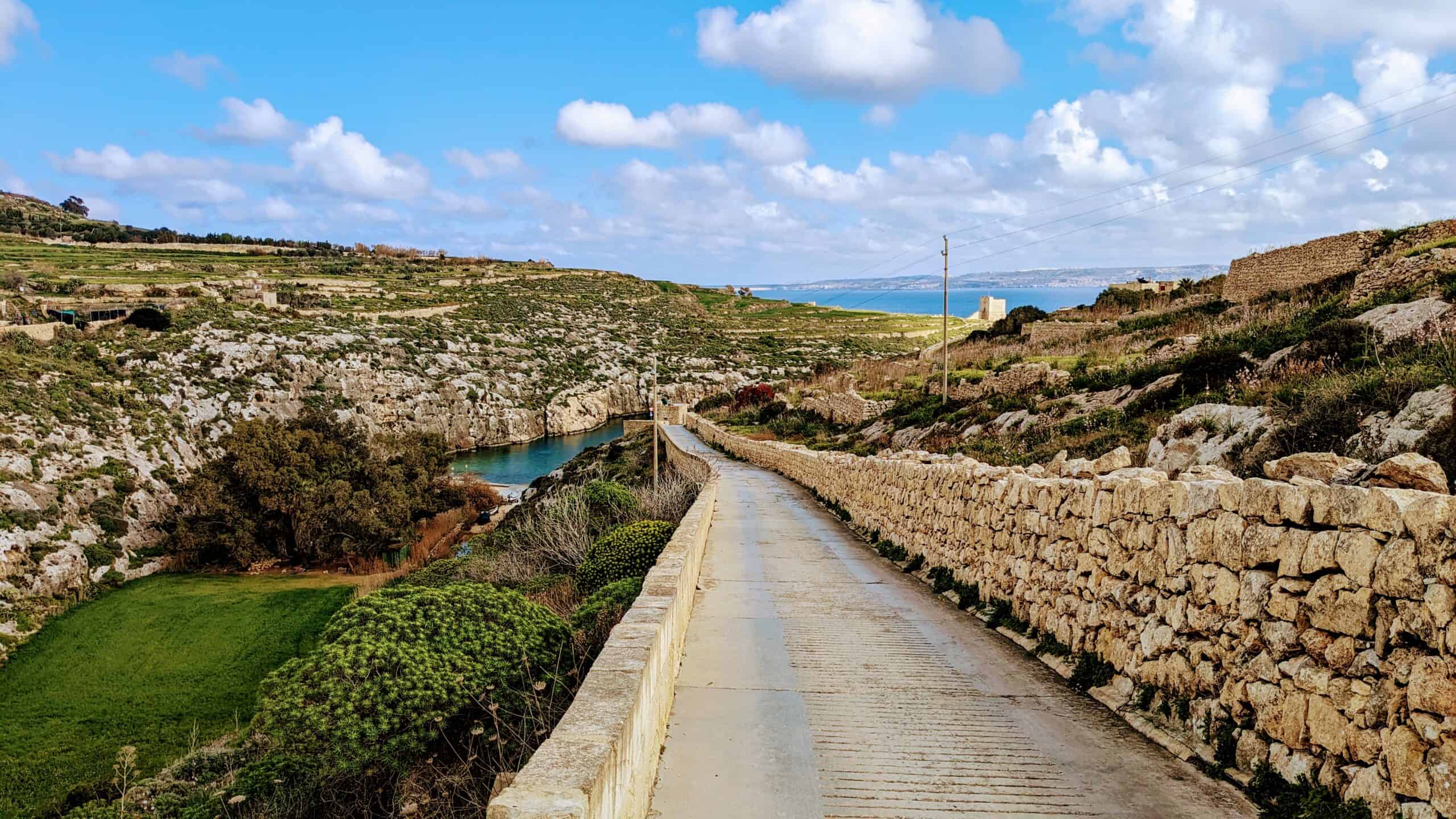 A narrow, scenic road bordered by stone walls winds through Malta's verdant landscape towards a distant body of water. The sky is partly cloudy, and the terrain features rolling hills and lush vegetation, making it a must-see in any Malta travel guide of things to do on the island.