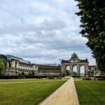 In Brussels, a wide, grassy park with a central dirt path leads to a grand archway flanked by historic buildings. Lush trees line the path under an overcast sky, creating a scene both serene and expansive.