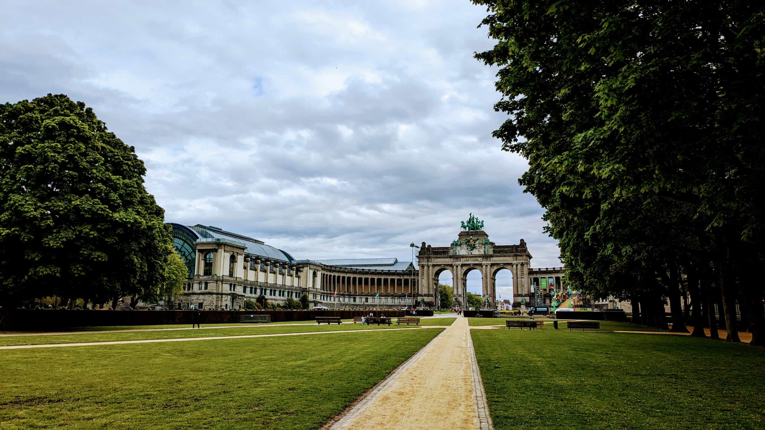 In Brussels, a wide, grassy park with a central dirt path leads to a grand archway flanked by historic buildings. Lush trees line the path under an overcast sky, creating a scene both serene and expansive.