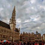 A bustling crowd with umbrellas gathers in the historic Grand Place of Brussels, surrounded by ornate Gothic buildings, including the towering Town Hall. The overcast sky is filled with dramatic clouds, perfectly capturing the enchanting atmosphere of Belgium's capital city.