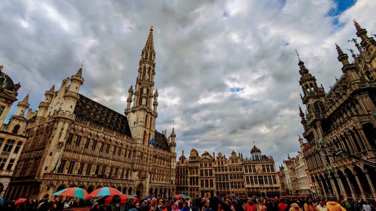 A bustling crowd with umbrellas gathers in the historic Grand Place of Brussels, surrounded by ornate Gothic buildings, including the towering Town Hall. The overcast sky is filled with dramatic clouds, perfectly capturing the enchanting atmosphere of Belgium's capital city.