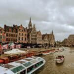 The canal in the European city of Ghent is flanked by historic buildings with ornate facades. Several boats with people glide along the water beneath a cloudy sky, while a blend of old and modern architecture graces the waterfront.