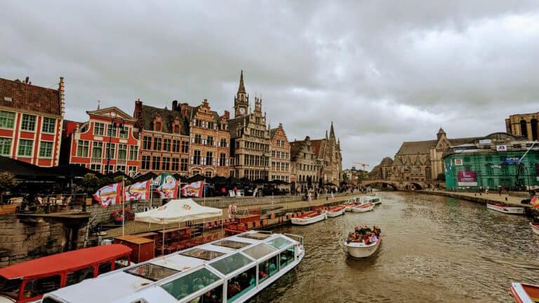The canal in the European city of Ghent is flanked by historic buildings with ornate facades. Several boats with people glide along the water beneath a cloudy sky, while a blend of old and modern architecture graces the waterfront.