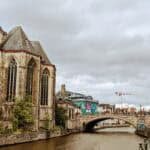A scenic view of Ghent reveals a European city with a canal in the foreground. On the left, a large historic building with pointed roofs and arched windows stands proudly. In the background, a bridge spans the canal under a cloudy sky as pedestrians leisurely stroll along the waterway.