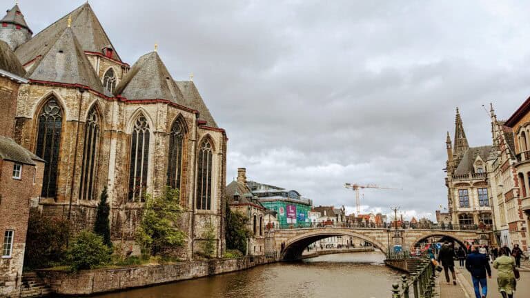 A scenic view of Ghent reveals a European city with a canal in the foreground. On the left, a large historic building with pointed roofs and arched windows stands proudly. In the background, a bridge spans the canal under a cloudy sky as pedestrians leisurely stroll along the waterway.
