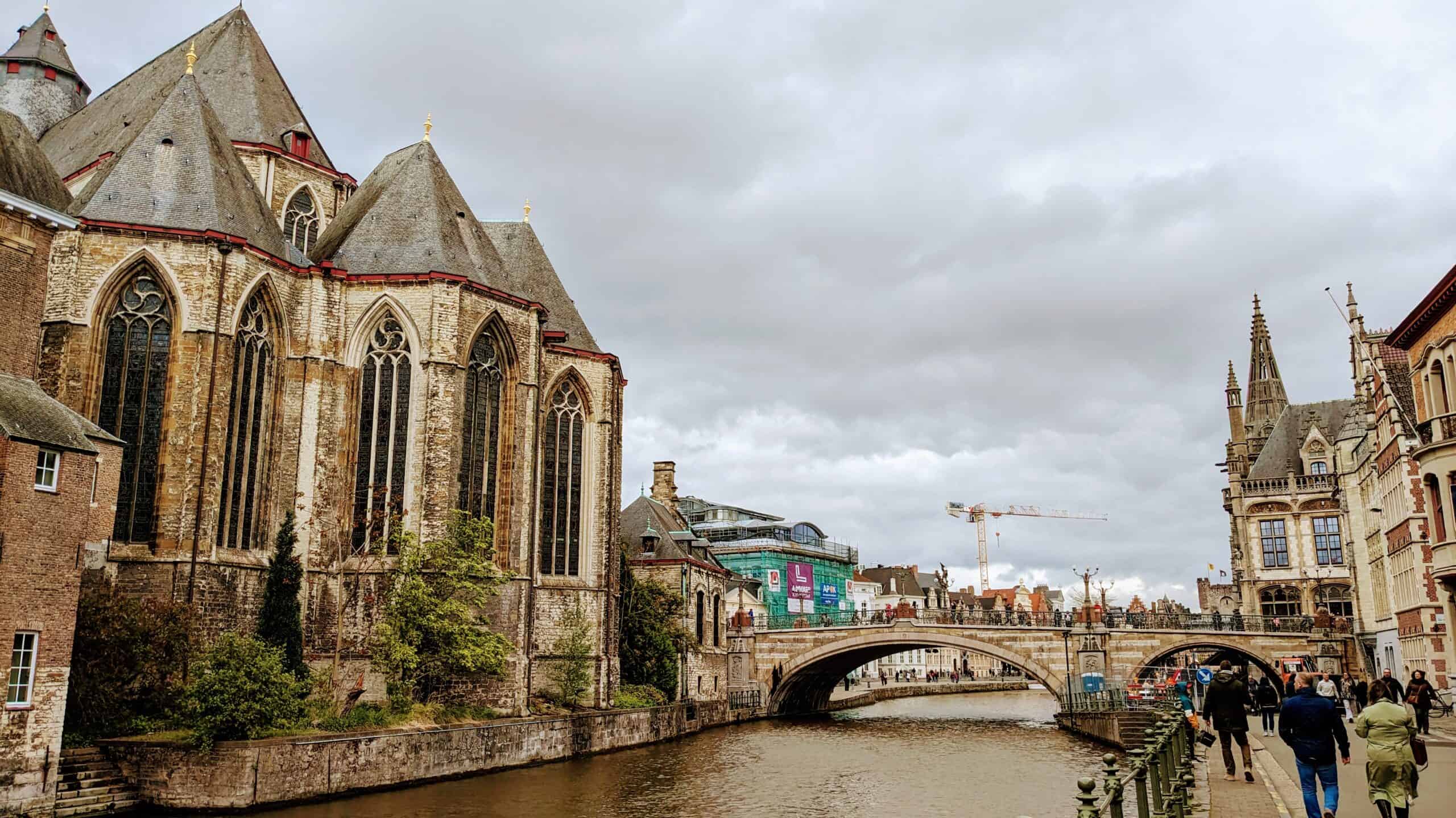 A scenic view of Ghent reveals a European city with a canal in the foreground. On the left, a large historic building with pointed roofs and arched windows stands proudly. In the background, a bridge spans the canal under a cloudy sky as pedestrians leisurely stroll along the waterway.