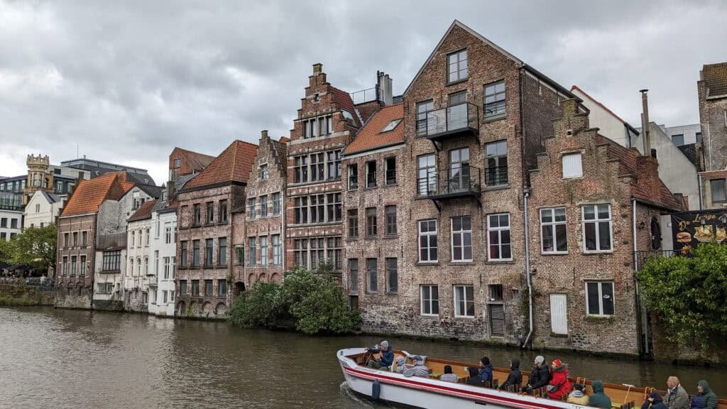 A canal boat carrying passengers glides through Ghent, past historic brick buildings with stepped gables lining the waterfront. The overcast sky enhances the moody atmosphere of this charming Belgian city.