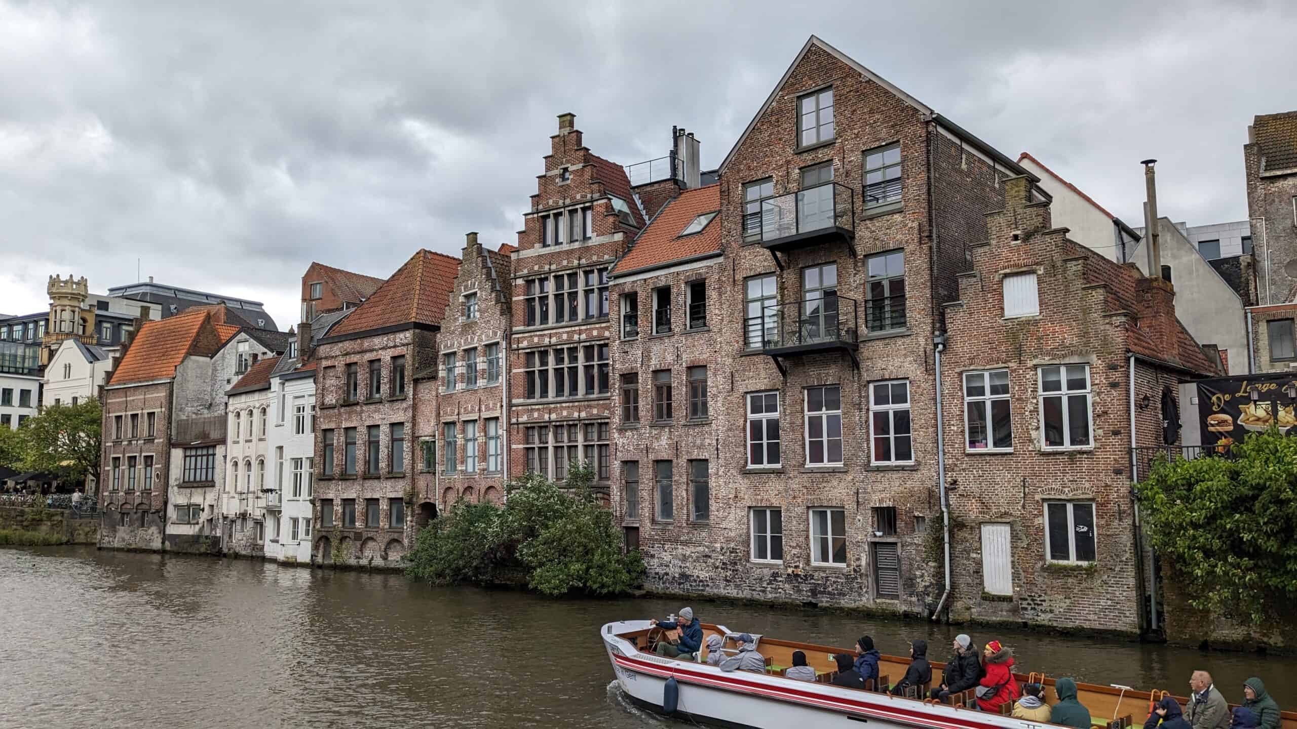 A canal boat carrying passengers glides through Ghent, past historic brick buildings with stepped gables lining the waterfront. The overcast sky enhances the moody atmosphere of this charming Belgian city.