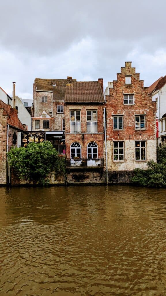 Historic brick buildings line a canal in Ghent, showcasing arched windows and stepped gables. The scene is mirrored in the brownish water under a cloudy sky, with lush vegetation accentuating the base of these charming structures.