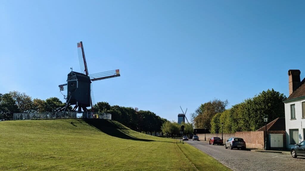 Two windmills sit on a lush green hill, with a road and some cars to the right. The sky is deep blue.