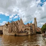 A picturesque view of Bruges, Belgium, featuring historic brick buildings along a canal under a partly cloudy sky. People are on a boat in the water, while others walk along the scenic waterfront, capturing the charm of this medieval city and checking off things to do.