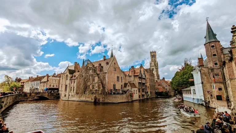 A picturesque view of Bruges, Belgium, featuring historic brick buildings along a canal under a partly cloudy sky. People are on a boat in the water, while others walk along the scenic waterfront, capturing the charm of this medieval city and checking off things to do.