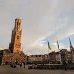 A scenic view of Bruges' city square at sunset, featuring the tall, historic Belfry tower. Several flags line this iconic Belgium location, and bicycles are parked in the foreground. The sky is partly cloudy, casting a warm glow over one of the best things to do in Europe.