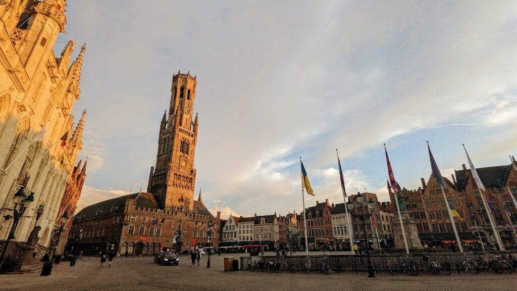 A scenic view of Bruges' Markt Square at sunset captures the Belfry tower prominently against a partly cloudy sky. This Belgian gem is lined with historic buildings and colorful flags, with bicycles parked nearby and few people strolling about, making it a must-see on your list of things to do.