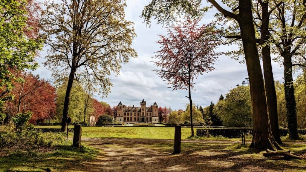 A grand castle with multiple towers rises in the distance, nestled amidst lush greenery and vibrant trees. This enchanting scene, reminiscent of Bruges' timeless charm, is framed by a rustic wooden fence and a dirt path under a partly cloudy sky—a must-see for anyone exploring Belgium.