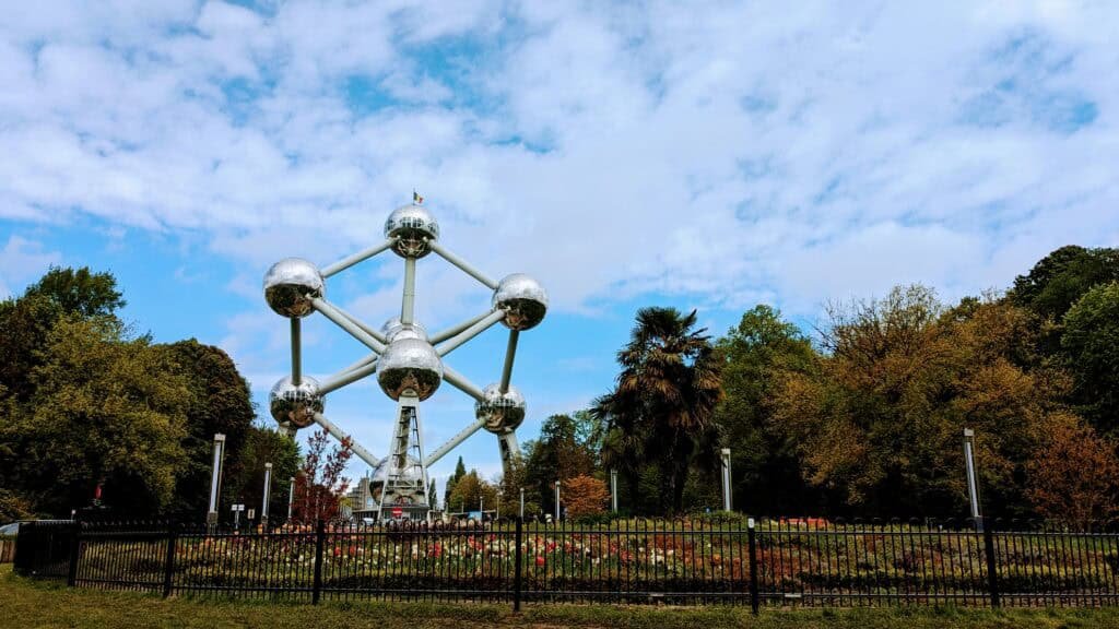 The image showcases the Atomium, a prominent metallic structure in Brussels, Belgium. Spherical nodes are linked by tubular steel beams, all set against a partly cloudy sky. Autumnal trees and a garden enrich the foreground, perfectly capturing the essence of Brussels' charm.