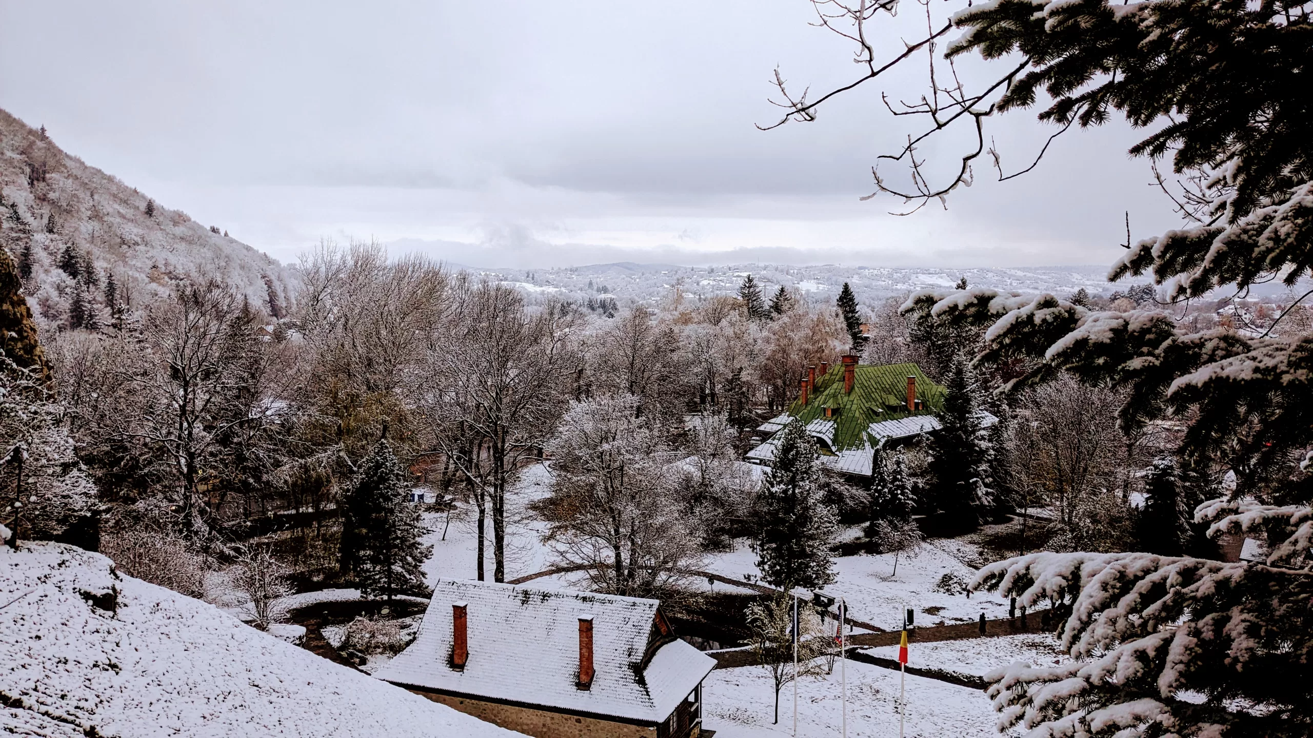 A snowy landscape resembling the outskirts of picturesque Bucharest features houses with snow-covered roofs nestled among bare trees. Evergreen branches frame the foreground, while rolling hills under a cloudy sky create a serene winter scene reminiscent of things to do in Bucharest during winter.