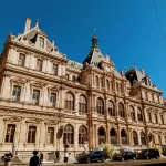 A grand historic building with intricate architecture, featuring tall windows and ornate details, set against a clear blue sky. People and cars bustling in the foreground suggest it as one of the must-see things to do in Lyon.