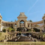 The Palais Longchamp in Marseille, with its ornate fountain and columns framed by greenery against a blue sky.