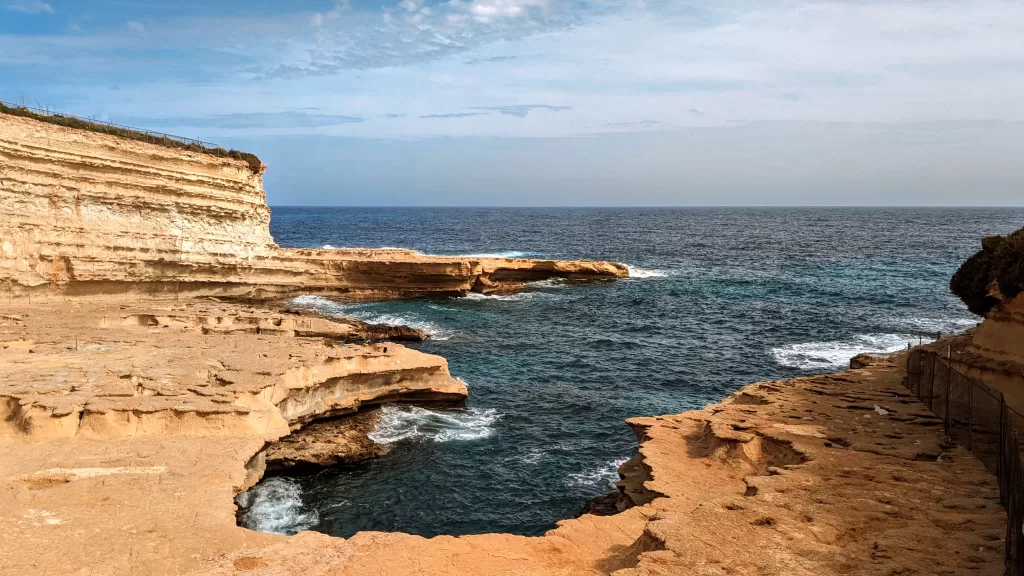 A limestone cove cut into the cliff side, with a dark blue sea and froth from the sea crashing on the rocks.