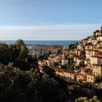 A hillside cityscape in Nice boasts colourful buildings nestled among trees, with views of the distant ocean under a clear blue sky.