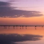 A tranquil sunset over a calm body of water in the south of France, with silhouettes of flamingos wading. The sky transitions from orange to pink, mirrored in the water's reflection, with clouds partially covering the horizon.