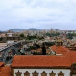 A stunning cityscape view of Porto, Portugal, showcases the iconic Dom Luís I Bridge gracefully arching over the Douro River. Historic buildings with red-tiled roofs fill the foreground, while modern structures rise in the background beneath a cloudy sky, capturing Porto's unique blend of old and new.