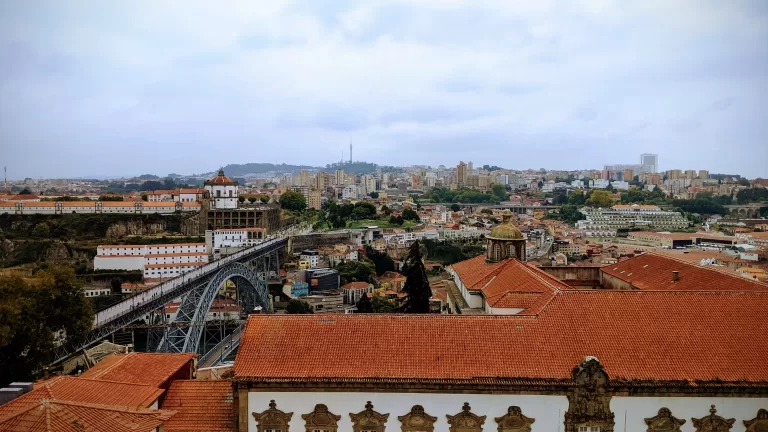 A stunning cityscape view of Porto, Portugal, showcases the iconic Dom Luís I Bridge gracefully arching over the Douro River. Historic buildings with red-tiled roofs fill the foreground, while modern structures rise in the background beneath a cloudy sky, capturing Porto's unique blend of old and new.