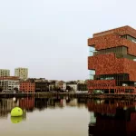 Modern red brick building reflecting in water with cityscape background, showcasing innovative architecture and urban scenery.