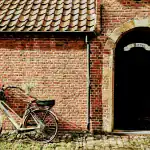 Vintage bicycle with basket parked against a rustic brick wall near a black door with a stone archway.