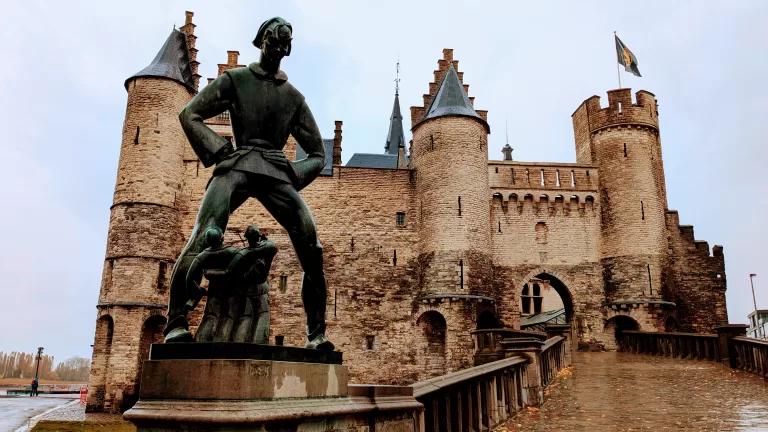 A modern bronze statue of a giant and some men stand in front of a grand, medieval castle