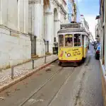 A vintage yellow tram glides down a narrow street in Lisbon, Portugal, a must-see among things to do in Lisbon. It passes historic buildings with stone facades as a woman with a stroller walks on the cobbled pavement, while another tram is visible on the opposite side.