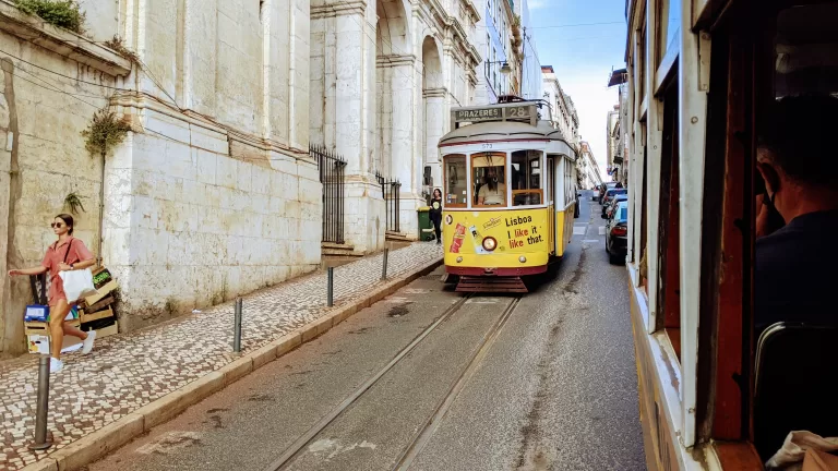 A vintage yellow tram glides down a narrow street in Lisbon, Portugal, a must-see among things to do in Lisbon. It passes historic buildings with stone facades as a woman with a stroller walks on the cobblestone sidewalk, while another tram is visible on the opposite side.