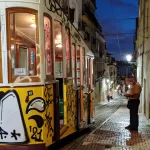A colourful tram covered in graffiti travels down a narrow, cobblestone street lined with old buildings at dusk. A man stands beside the tram, looking at his phone. Streetlights illuminate the scene.