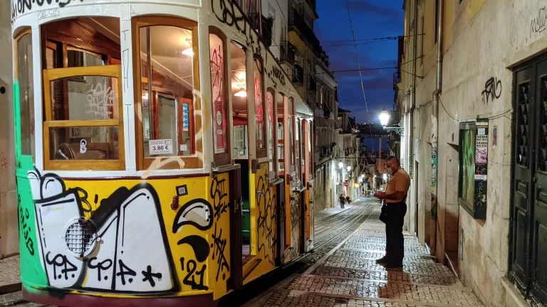 A colorful tram covered in graffiti travels down a narrow, cobblestone street lined with old buildings at dusk. A man stands beside the tram, looking at his phone. Streetlights illuminate the scene.