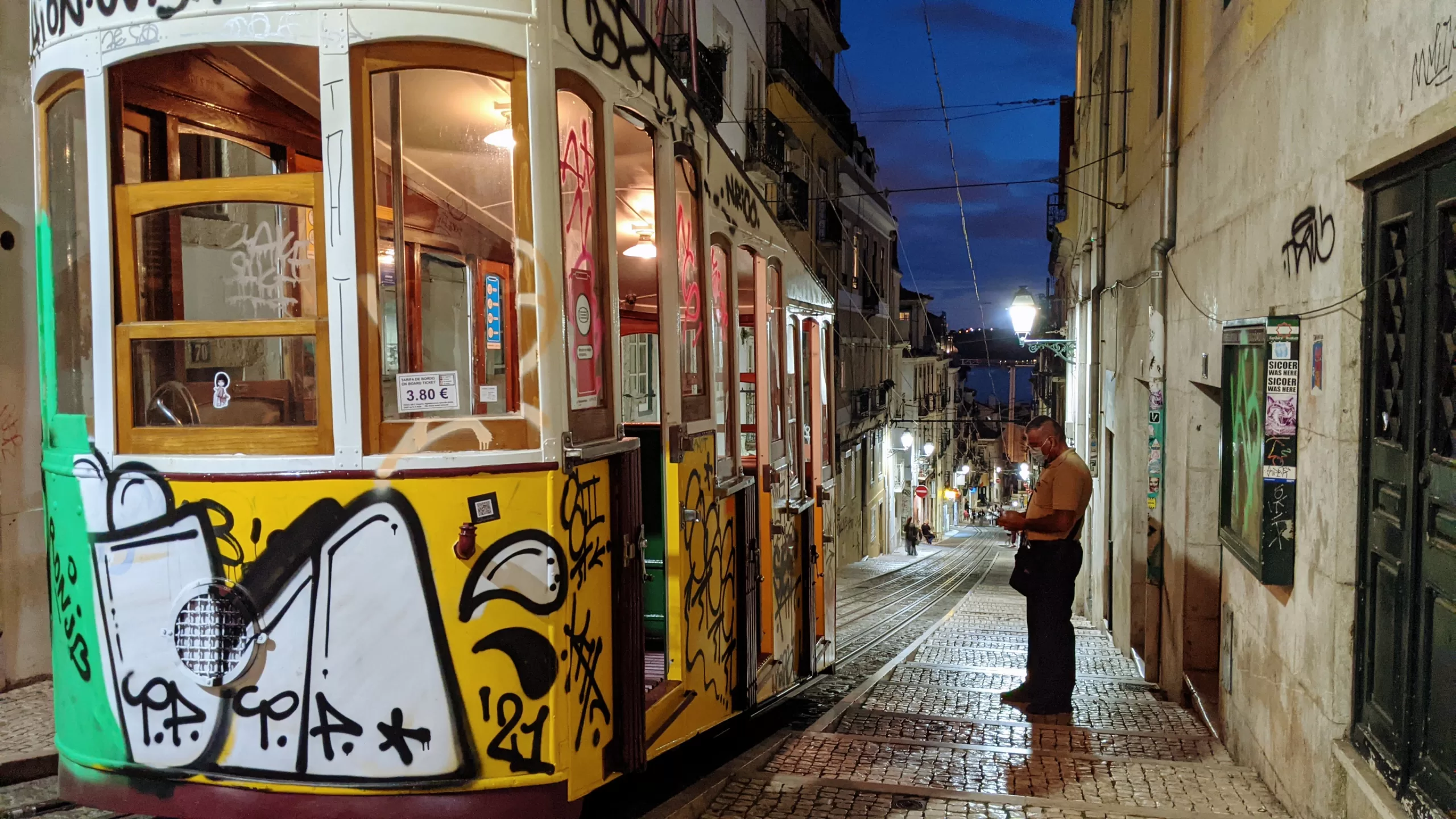 A colorful tram covered in graffiti travels down a narrow, cobblestone street lined with old buildings at dusk. A man stands beside the tram, looking at his phone. Streetlights illuminate the scene.