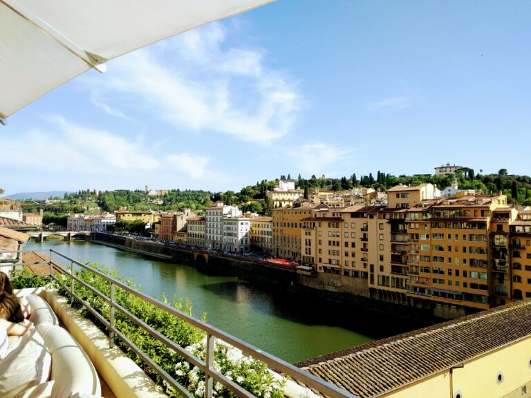 The green Alto river stretches by surrounded by tall, pastel coloured buildings. There is an umbrella on the top left from the roof top bar where this is taken from. There is a blue, slightly cloudy sky.