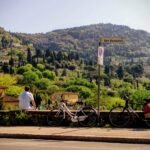 The green, tree-filled hills of Tuscany rise into the distance. There is a road in the foreground with two bikes and a person sitting on a wall, looking at the view.
