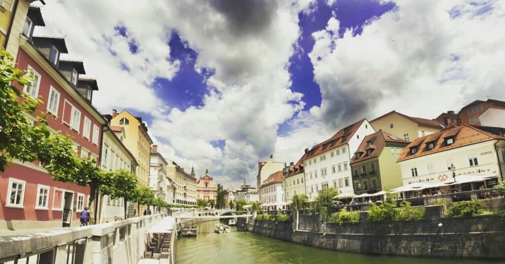 Scenic view of colorful riverside buildings in Ljubljana, showcasing vibrant architecture under dramatic skies.