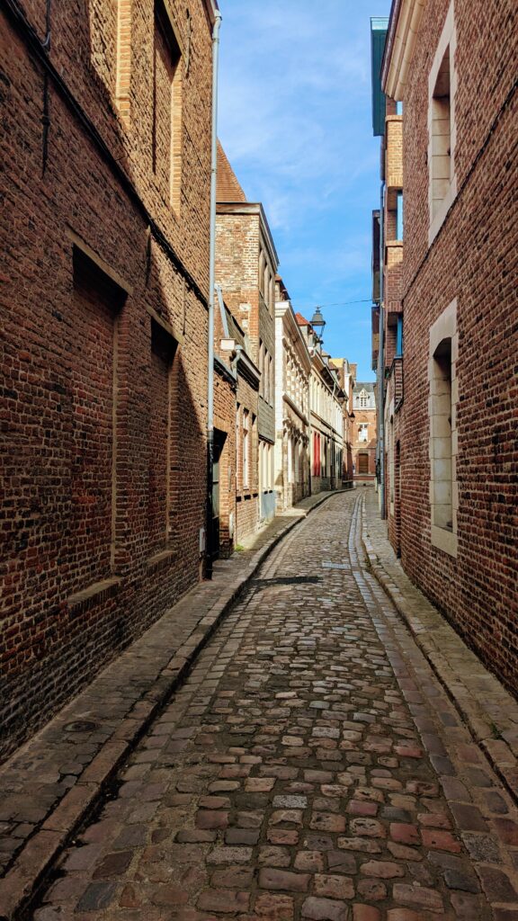 Old cobblestone alleyway in historic European town, narrow street lined with brick buildings, charming and picturesque with clear blue sky, perfect for travel photography and exploring off-the-beaten-path destinations.