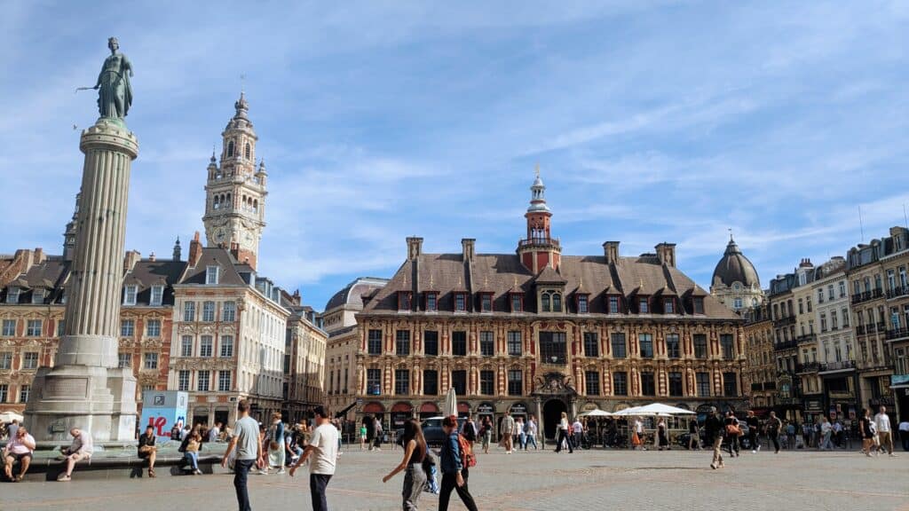 Historic square with Renaissance architecture in Lille, France, featuring a statue, clock tower, and lively crowd under a bright blue sky. The old town will form a big part of your perfect weekend in Lille.