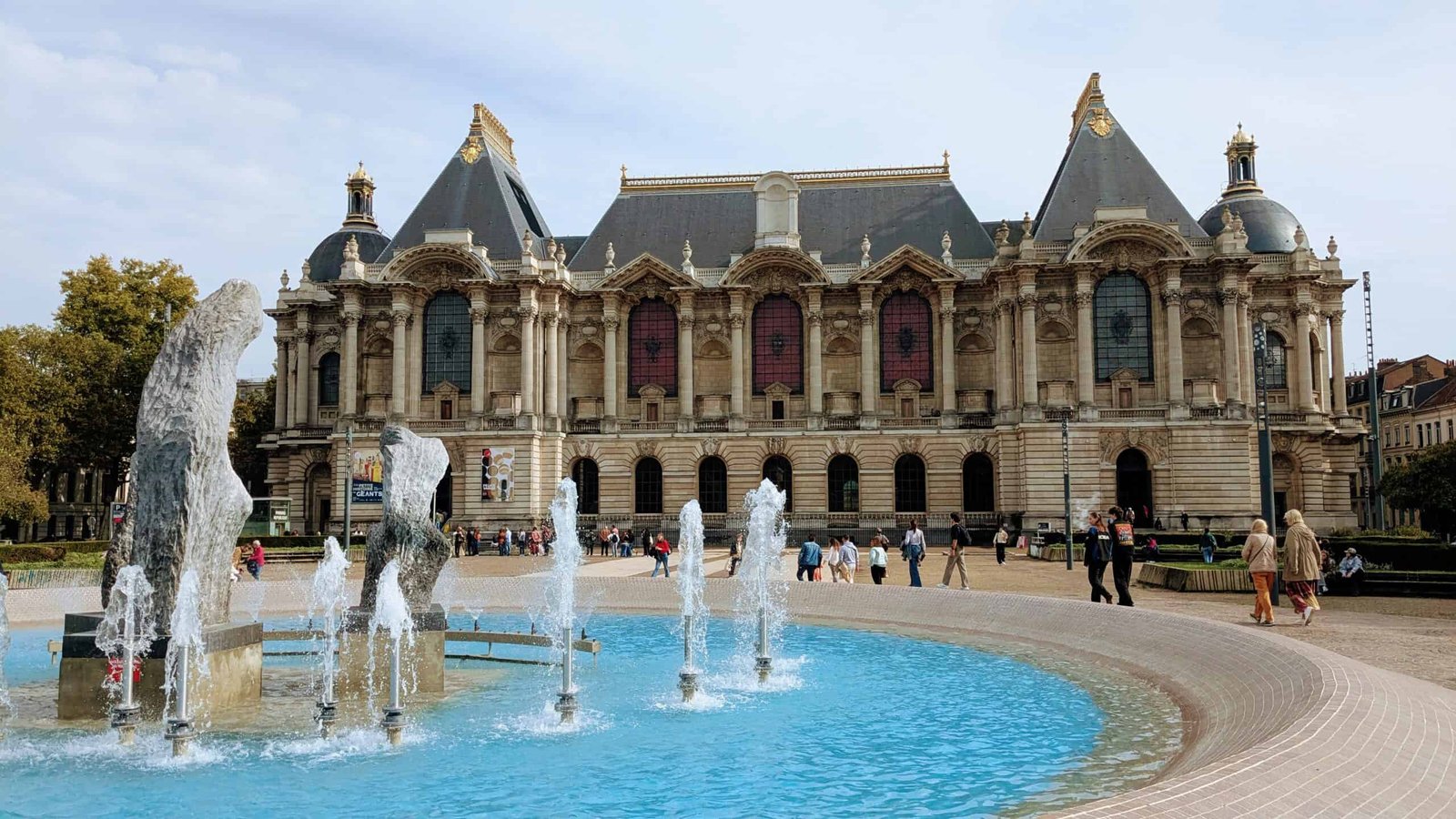 Intricate historic theatre building with fountain in foreground, surrounded by visitors enjoying a sunny day.