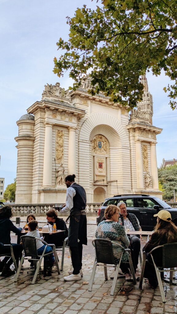 Historic city square with outdoor café seating in front of a grand ornate monument and lush green trees, perfect for travellers exploring cultural landmarks on a trip.