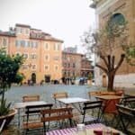An outdoor seating area in a historic European square with wooden tables and chairs, potted plants, and a large tree, surrounded by colourful old buildings and a church.