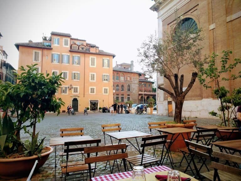 An outdoor seating area in a historic European square with wooden tables and chairs, potted plants, and a large tree, surrounded by colourful old buildings and a church.