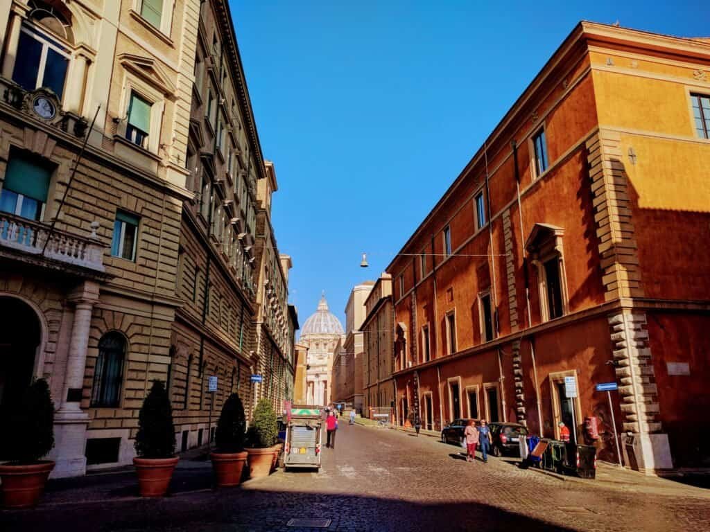 An urban view of a charming Roman street featuring classic architecture, cobblestone pavement, and the iconic dome of St. Peter's Basilica in the distance, capturing the essence of travel in Italy.