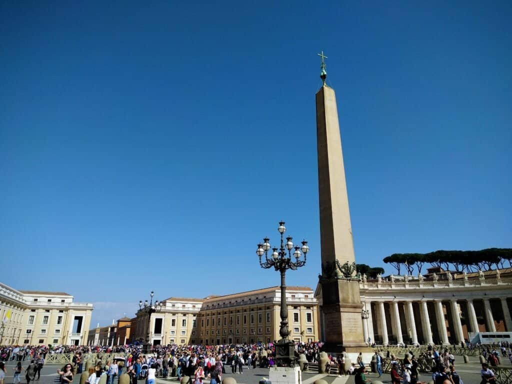A bustling scene in St. Peter's Square with tourists and visitors gathering around the historic obelisk under a clear blue sky.